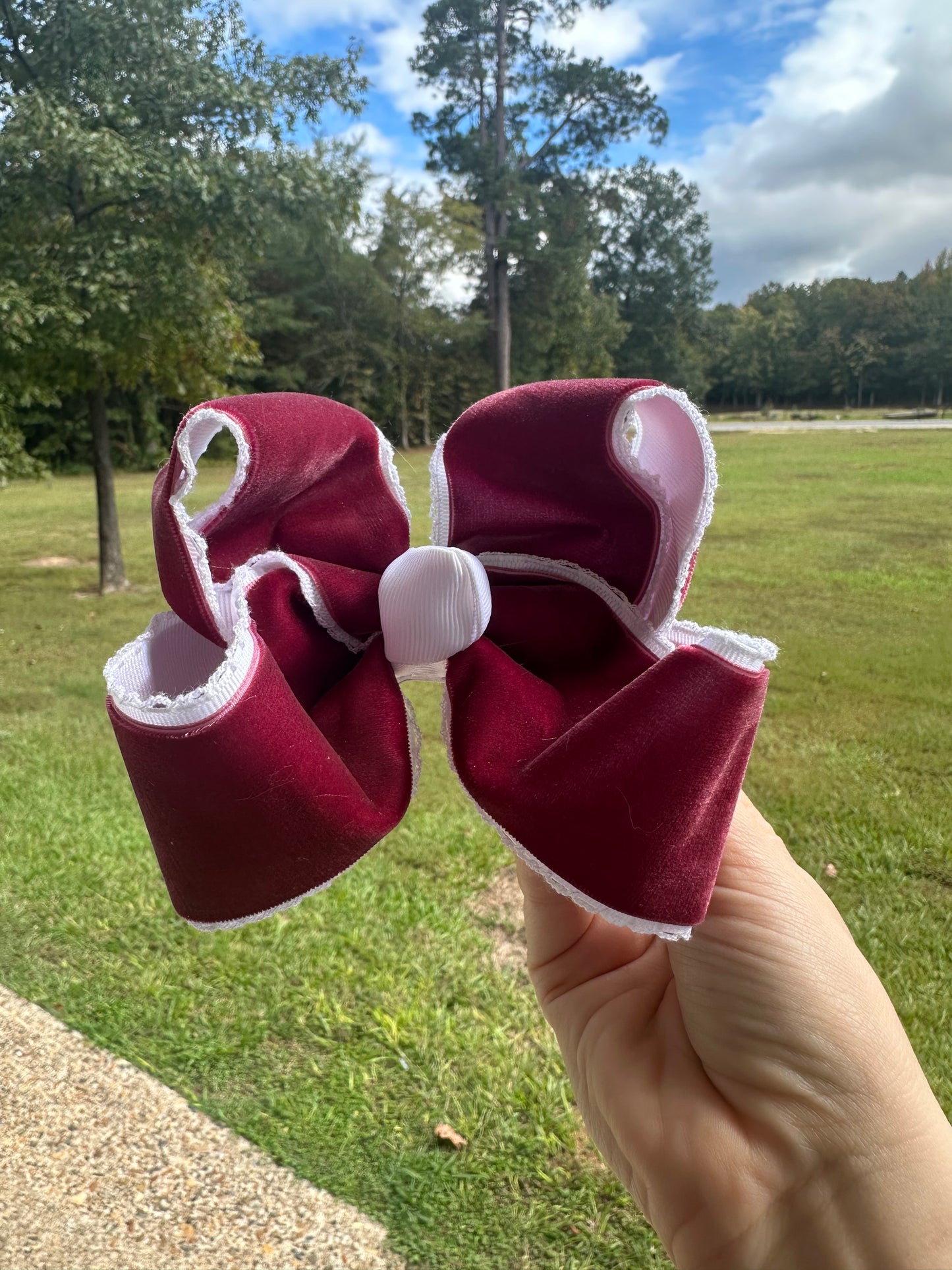 Deep red velvet hair bow with white lace trim and white center wrap, held outdoors against a grassy background and blue sky